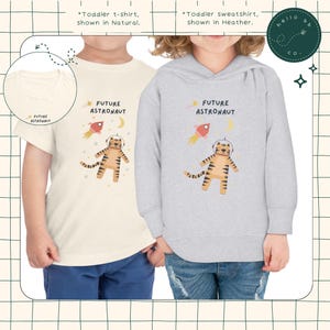 Peut inclure: Deux v&ecirc;tements pour enfants repr&eacute;sentant un tigre portant un casque d'astronaute et une fus&eacute;e. Le t-shirt est de couleur cr&egrave;me avec le texte "FUTURE ASTRONAUT" et le sweat-shirt est gris chin&eacute; avec le m&ecirc;me texte.