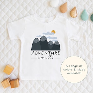 Peut inclure: T-shirt blanc avec un motif de montagnes, de nuages et d'un soleil. Le texte "ADVENTURE awaits" est imprimé sous le motif. Une gamme de couleurs et de tailles est disponible.