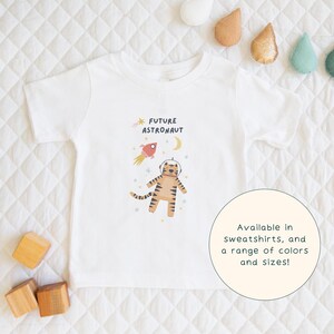 May include: White t-shirt with a graphic of a tiger wearing an astronaut helmet and a rocket ship. The text "FUTURE ASTRONAUT" is printed on the shirt.