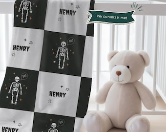 May include: A personalized baby blanket with a black and white checkered pattern. Each square features a skeleton graphic, the name "HENRY", and stars. A plush teddy bear sits beside the blanket. The blanket has the text "Personalize me!"