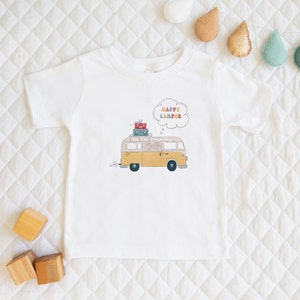 May include: White t-shirt with a yellow and brown vintage camper van illustration. The van has luggage on top and a thought bubble above it that says "Happy Camper".