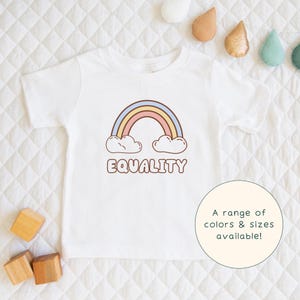 May include: White toddler t-shirt with a rainbow graphic and the word "EQUALITY" in brown letters. The rainbow has pastel colors and is above two white clouds. A circular graphic states "A range of colors & sizes available!"