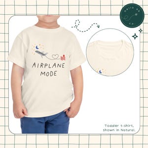 May include: A white toddler t-shirt featuring a graphic of an aeroplane flying over a heart and a house. The text "AIRPLANE MODE" is printed below the graphic. The shirt is shown in the colour "Natural".