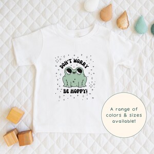 May include: White short-sleeved t-shirt with a green frog illustration and the text "Don't worry be hoppy!" printed on the front.