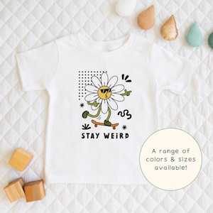 May include: White toddler t-shirt featuring a cartoon daisy wearing sunglasses and riding a skateboard. The daisy has a yellow center and white petals. The text "STAY WEIRD" is printed below the daisy in black. The shirt is on a white quilted surface.