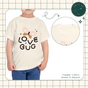 May include: A toddler wearing a white t-shirt with a butterfly graphic and the text "LOVE BUG" in brown and black lettering. The shirt is shown in the color Natural.