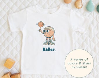 May include: White t-shirt featuring a cartoon basketball player dribbling a ball. The text "Baller." is printed below the image. A range of colours and sizes are available.