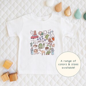 May include: White t-shirt with a colorful illustration of various Eid al-Fitr symbols, including a mosque, crescent moon, stars, and a camel. The text "Happy Eid" is written in the center of the design.