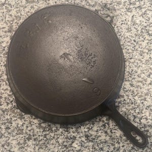 May include: A well-worn, cast iron skillet with a long handle and a circular cooking surface. The underside of the pan is visible, showing the number "9" and other markings. The pan has a dark, matte finish and is designed for stovetop cooking.