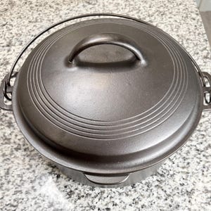 May include: A round, dark gray cast iron Dutch oven with a matching lid. The lid features three concentric circular ridges and a central arched handle. A thin metal wire bail handle is attached to the side lugs of the pot.