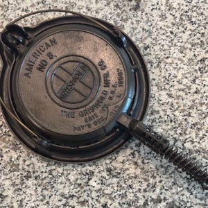 Pode incluir: Caneca de cerâmica branca com uma alça preta e o texto "But First, Coffee" em estilo manuscrito. A caneca está sobre uma superfície branca. Adequada para bebidas quentes.