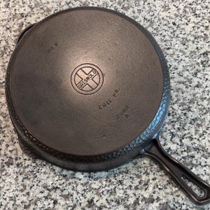 May include: A cast iron Griswold No. 8 skillet, viewed from the bottom. The pan is dark gray with a textured surface and a long handle. The Griswold logo and "ERIE PA. 2008 A" are stamped on the base. The skillet is designed for cooking.