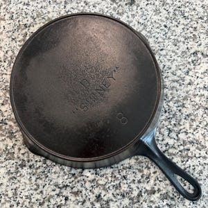 May include: A black cast iron skillet with a long handle and a loop for hanging. The bottom of the pan has the stamped text "D SIDNEY" and the number "8". The pan is round and has a textured surface.