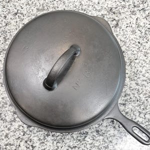 May include: A round, black cast iron frying pan with a matching lid. The lid features a central handle and the embossed text 'No. 8'. The pan has a single long handle with a hole at the end and a small pour spout visible on the side of the rim.