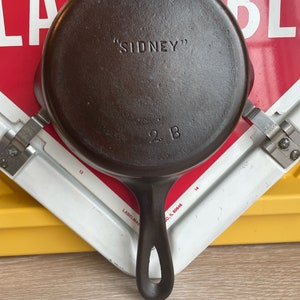 Könnte beinhalten: Eine gusseiserne Bratpfanne mit einem schwarzen Griff. Die Bratpfanne ist mit "SIDNEY" und "2 B" beschriftet.