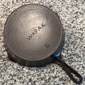 Pode incluir: Uma frigideira de ferro fundido com acabamento cinza escuro. A palavra "WAPAK" está gravada na base, juntamente com o número "6". A frigideira tem uma alça e está sobre uma superfície salpicada.