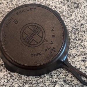 Op de afbeelding: Een donkerbruine Griswold 8 gietijzeren koekenpan, van onderaf gezien. De pan heeft de woorden "CAST IRON SKILLET 8" en "GRISWOLD ERIE PA. U.S.A." in reliëf op het oppervlak. De handgreep heeft een gat om op te hangen.