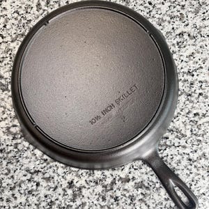 May include: A round, dark gray cast iron skillet viewed from the underside. The base features embossed text that reads 10 1/2 INCH SKILLET and the number 2. The skillet has a single handle with an elongated hole at the end for hanging storage.