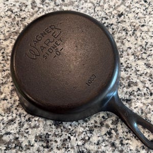 以下が含まれることがあります： 片手鍋の焦げ茶色の鋳鉄製Wagner Ware。鍋底にはWagner Wareのロゴと「Sidney -0-」がエンボス加工されています。数字の「1053」も確認できます。鍋はまだらの表面に置かれています。