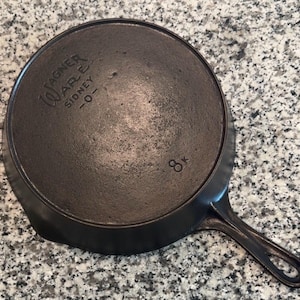 May include: A black cast iron skillet with a handle, on a speckled surface. The underside of the pan has the stamped text "WAGNER WARE SIDNEY -0-" and the number "8k".