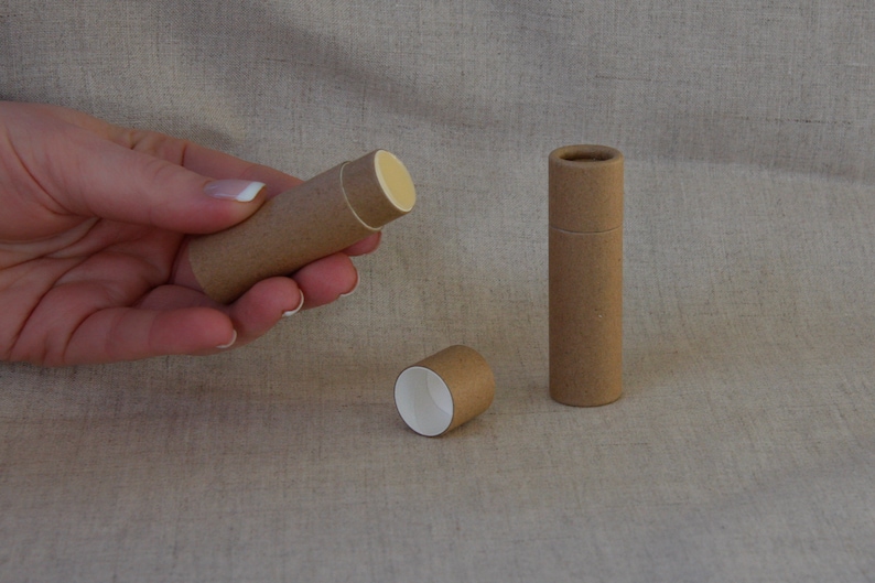 May include: Three brown cardboard tubes, one with a white interior, one with a white interior and a yellow lip balm inside, and one with a white interior and a yellow lip balm inside. The tubes are on a beige fabric surface.
