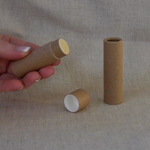 May include: Three brown cardboard tubes, one with a white interior, one with a white interior and a yellow lip balm inside, and one with a white interior and a yellow lip balm inside. The tubes are on a beige fabric surface.