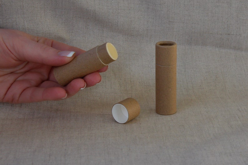 May include: Three brown cardboard tubes, one with a white interior, one with a white interior and a yellow lip balm inside, and one with a white interior and a yellow lip balm inside. The tubes are on a beige fabric surface.