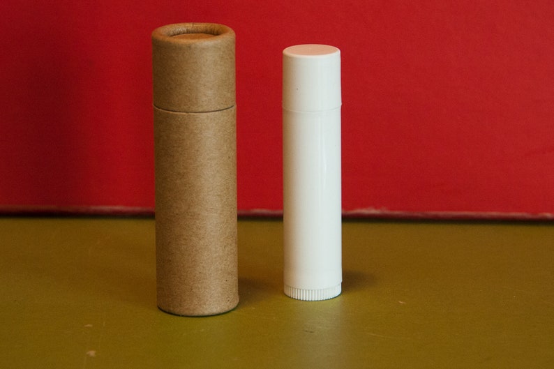 May include: Two tubes of lip balm, one brown and one white, on a green surface.
