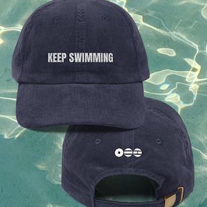 Puede incluir: Gorra de béisbol de pana azul marino con el texto "KEEP SWIMMING" bordado en blanco. La parte trasera de la gorra presenta un logotipo blanco y una hebilla ajustable dorada.