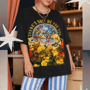 Puede incluir: Camiseta negra con el texto "THERE'S ONLY ME AND YOU" sobre una bola de discoteca y flores amarillas. La persona lleva gafas de sol blancas, collares dorados y pantalones a rayas azules y blancas. El fondo incluye una estantería con vasos y botellas.