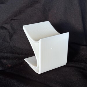 May include: A white, curved phone stand. The stand is made of a solid material and has a modern design. It features a curved base and a vertical support to hold a phone or tablet. The stand is designed to be stable and functional.