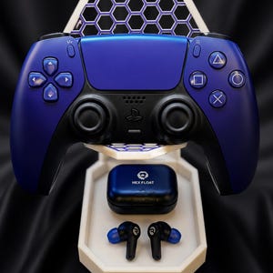 May include: A blue gaming controller rests on a white hexagonal stand featuring a honeycomb pattern. Below the controller, a black charging case labeled HEX FLOAT and a pair of matching black and blue wireless earbuds sit on the stand base.