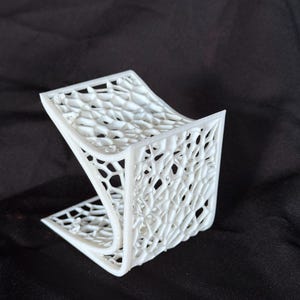 May include: A white, 3D-printed phone stand with a unique, open-weave design. The stand is angled and has a curved base, providing a stable platform for a phone. The intricate pattern creates a modern, artistic aesthetic.