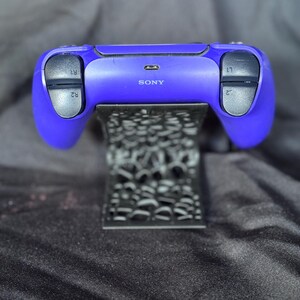 Może przedstawiać: Fioletowy kontroler Sony PlayStation 5 spoczywający na czarnym, dekoracyjnym stojaku. Kontroler ma czarne przyciski i spusty oznaczone R1, R2, L1 i L2. Stojak ma wz&oacute;r.