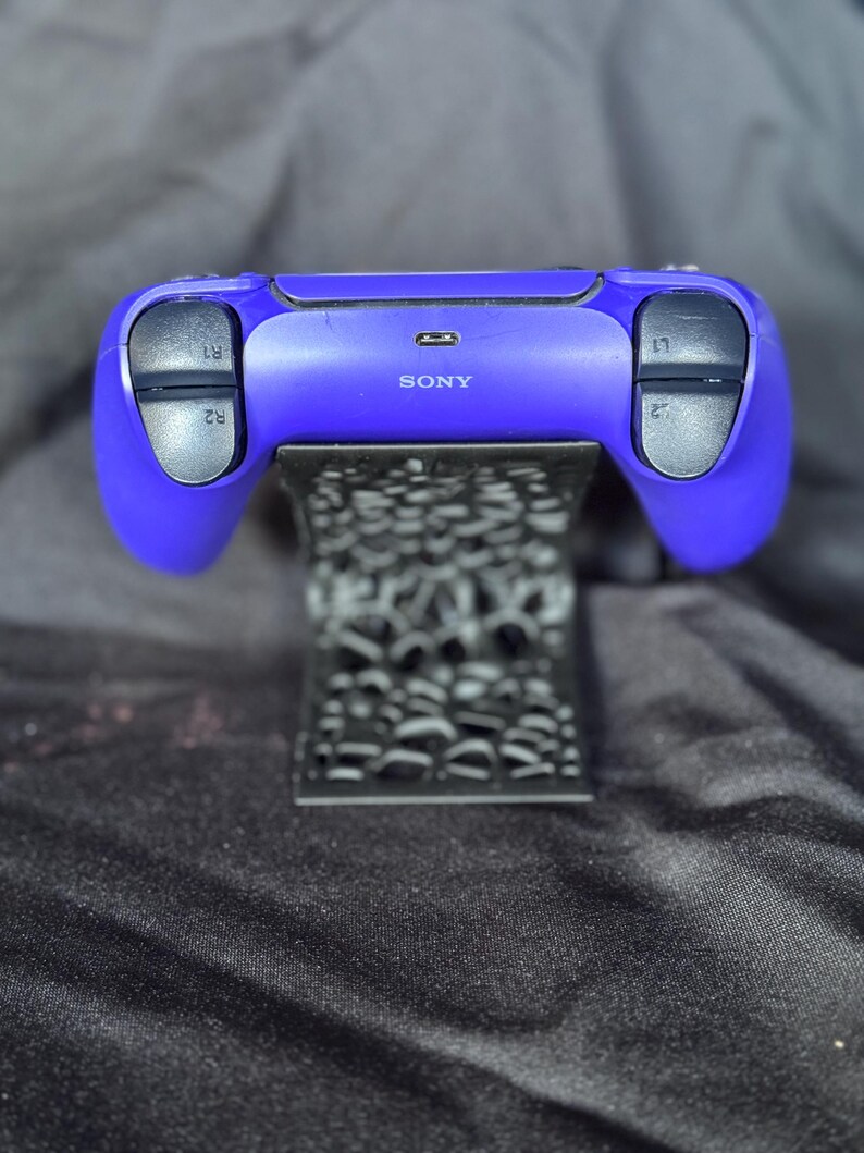 Może przedstawiać: Fioletowy kontroler Sony PlayStation 5 spoczywający na czarnym, dekoracyjnym stojaku. Kontroler ma czarne przyciski i spusty oznaczone R1, R2, L1 i L2. Stojak ma wz&oacute;r.