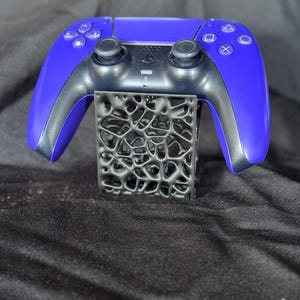 Może przedstawiać: Niebiesko-czarny bezprzewodowy kontroler do gier spoczywający na czarnym stojaku. Kontroler ma symetryczny kształt z przyciskami i joystickami. Stojak ma unikalny, ażurowy wz&oacute;r.