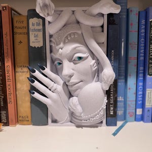 May include: White bookends shaped like a woman's head with snakes for hair. The woman has blue eyes and is holding a snake in her hand. The bookends are on a bookshelf with other books.