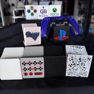 May include: A collection of 3D-printed gaming accessories, including controller stands and decorative blocks. The image features a blue and black PlayStation controller on a stand with the PlayStation logo. Other blocks display game controller button layouts and logos.