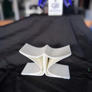 May include: A white, abstract, folded paper sculpture with curved edges, displayed on a black surface. The sculpture has a unique, open design. In the background, a blue and black object is visible.