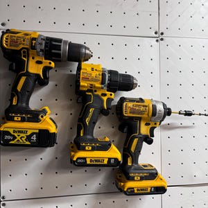 Op de afbeelding: Drie gele en zwarte DeWalt-boormachines met verschillende batterijgroottes hangen aan een witte pegboard-wand. De boormachines zijn gemarkeerd met "20V" en hebben verschillende ampère-uur ratings.