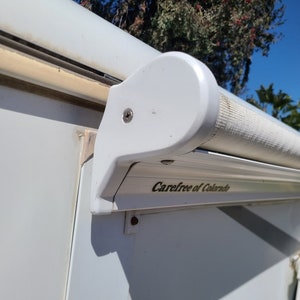 Pode incluir: Close-up de um toldo branco de autocaravana com o texto "Carefree of Colorado" impresso na lateral. O toldo está enrolado e montado na lateral da autocaravana. O céu é azul com palmeiras ao fundo.