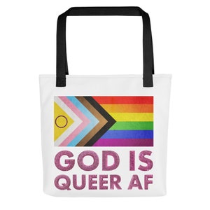 Może przedstawiać: Biała torba typu tote z czarnymi uchwytami. Torba przedstawia tęczową flagę dumy z żółtym kółkiem w lewym górnym rogu. Tekst "GOD IS QUEER AF" jest nadrukowany w różowym brokacie pod flagą.