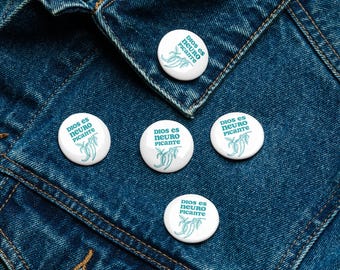 Può includere: Cinque bottoni rotondi bianchi con un design verde e il testo "DIOS es NEURO PICANTE" su uno sfondo di jeans blu.