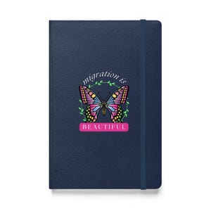 May include: A navy blue journal featuring a vibrant butterfly design. The butterfly is encircled by a green wreath, with the words "migration is BEAUTIFUL". The journal includes a matching elastic band.