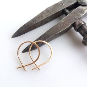 May include: A pair of delicate gold-colored hoop earrings with a simple, minimalist design. The earrings are positioned near a vintage metal tool, likely used in jewelry making. The background is a clean, bright white.