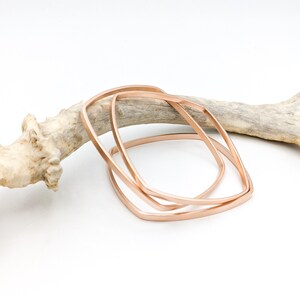 May include: Three rose gold square bangles stacked on top of each other. The bangles are on a light brown wooden surface.