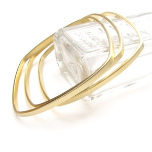 May include: Three gold-colored square bangles are stacked together, resting on a clear glass bottle. The bangles have a smooth, polished finish and a minimalist design. The background is white.