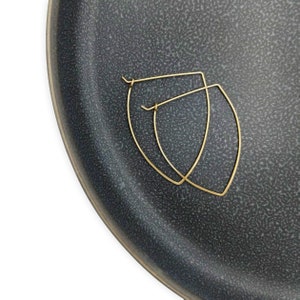 May include: A pair of gold-toned, geometric, leaf-shaped hoop earrings. The earrings are displayed on a black textured surface.
