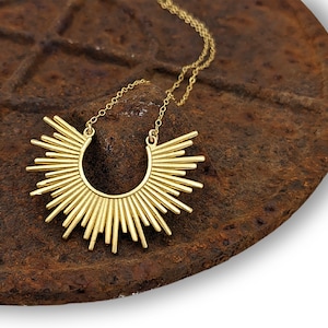 May include: A gold necklace with a sunburst pendant. The pendant is made of thin, vertical bars that radiate out from a central circle. The necklace is on a rusty metal surface.