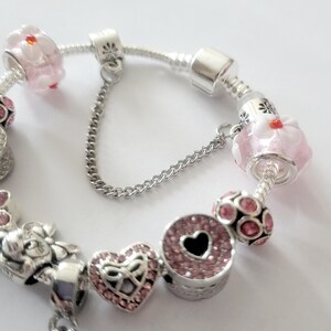 May include: A silver charm bracelet with various charms. Charms include pink flower beads, a heart-shaped charm with pink crystals, and a round charm with a black heart. The bracelet has a safety chain.